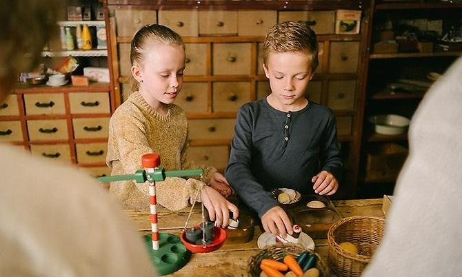 en flicka och en pojke står bakom disken av trä i handelsboden och leker affär. På disken står en korg med färgglada frukter i trä, en våg med mera. Bakom barnen syns en mäng lådor i rader i ljus färg med mörk träknopp