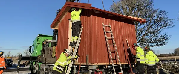 En friggebod står på en lastbil. Hantverkare arbetar med huset. Himmel i bakgrunden.